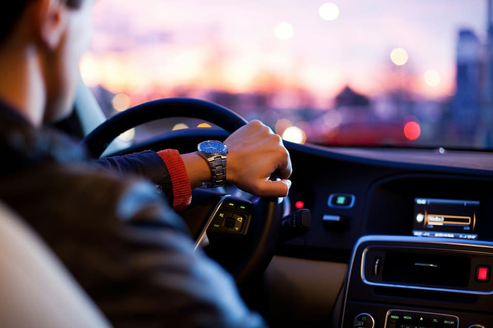 A driver at the wheel of a car at dusk, city lights blurred through the windscreen
